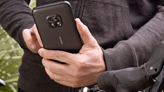 Um dos melhores telefones robustos, um Nokia Xr20 preto na mão de um homem. É simples com uma câmera na parte superior no meio.  o homem tem mãos brancas e está vestindo um moletom preto ao lado de uma bicicleta.