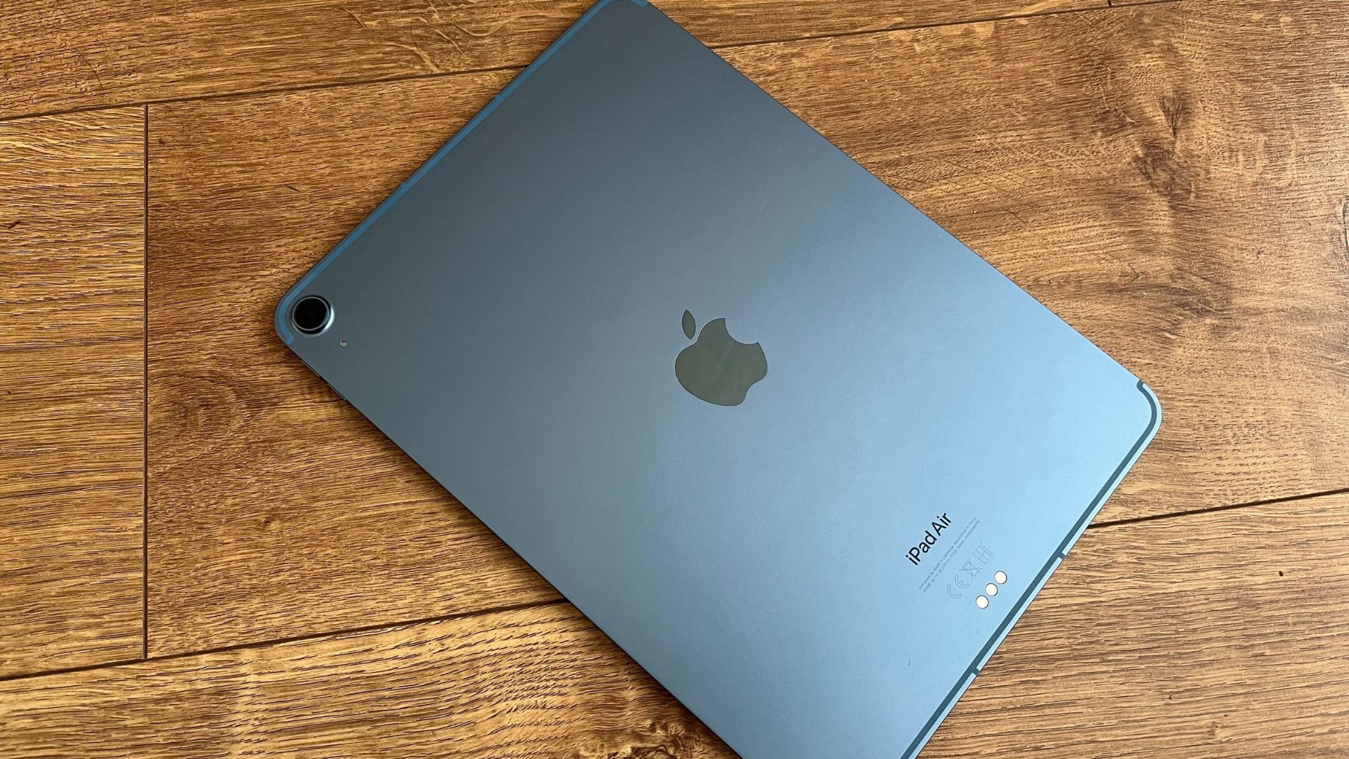 melhor-ipad-3 Um dos melhores iPads, o iPad Air, na cor preta, repousava sobre a tela sobre uma superfície de madeira.  Vemos sua parte traseira, mostrando o logotipo da Apple no centro e um pequeno anel de câmera única no canto superior direito.